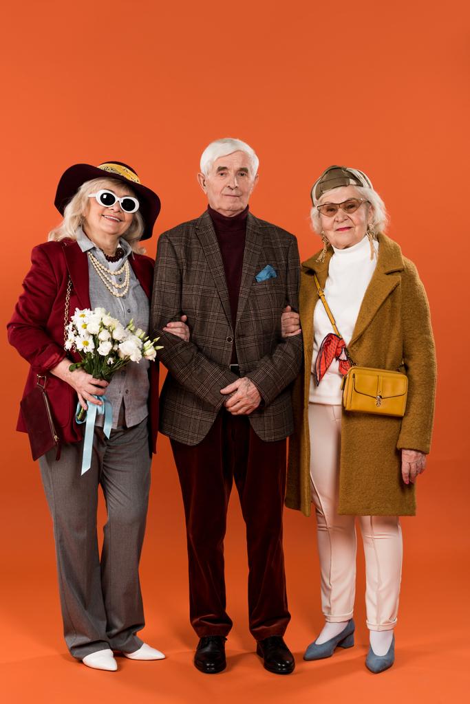 stijlvolle senior women permanent tussen gepensioneerde man en houden van bloemen op een oranje achtergrond - Foto, afbeelding