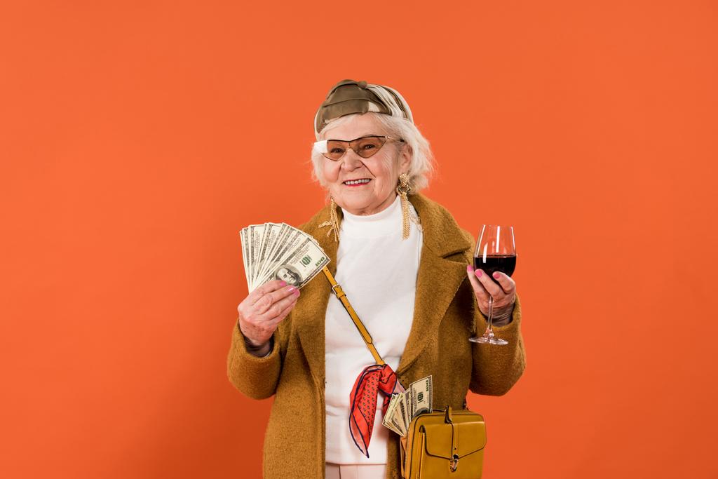 joyeuse femme âgée tenant de l'argent et un verre de vin dans les mains isolées sur orange
 - Photo, image