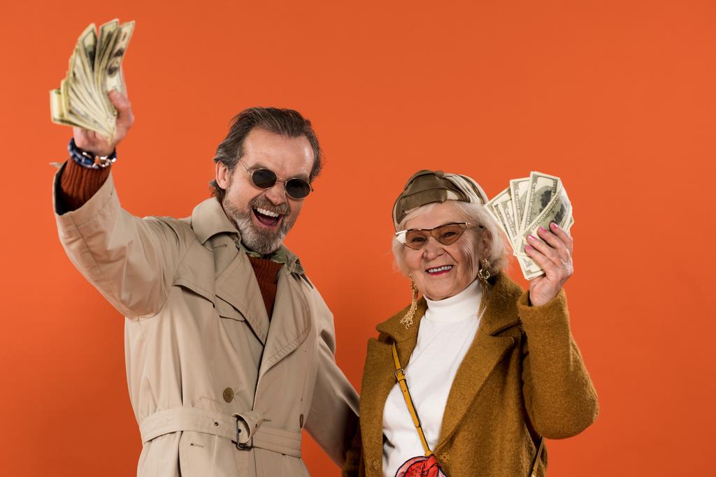 ευτυχισμένο ζευγάρι συνταξιούχων κρατώντας δολάριο τραπεζογραμματίων και χαμογελαστός απομονωμένες σε πορτοκαλί - Φωτογραφία, εικόνα