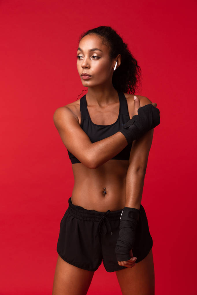 Foto de chica afroamericana de 20 años en ropa deportiva negra de pie con vendajes deportivos en sus manos aisladas sobre fondo rojo
 - Foto, Imagen