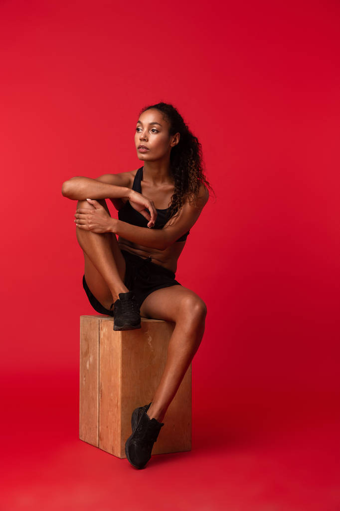 Ritratto di splendida donna afroamericana in abbigliamento sportivo nero seduta sulla scatola isolata su sfondo rosso
 - Foto, immagini