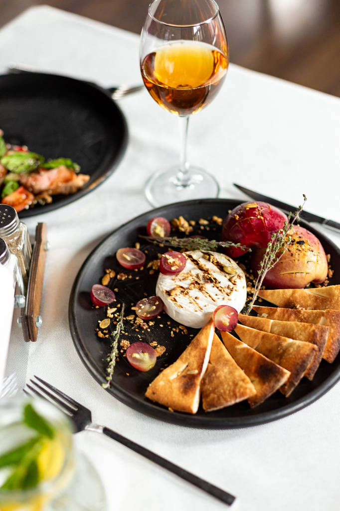 Grilled Camembert with baked apple and grapes with slices of bread tortilla, served on a black plate on a table with a white tablecloth, appliances and a glass of wine in a restaurant - Photo, Image