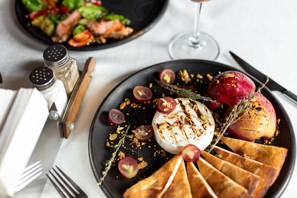 Camembert grelhado com maçã assada e uvas com fatias de tortilla de pão, servido em um prato preto em uma mesa com uma toalha de mesa branca, eletrodomésticos e um copo de vinho em um restaurante
 - Foto, Imagem