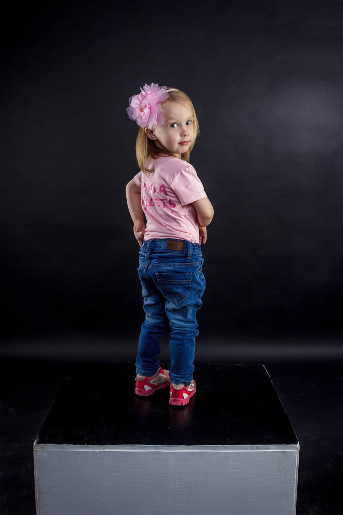 Портрет симпатичной европейской блондинки в джинсах и розовой футболке с надписью: "Папина гордость, мамина радость", на черном изолированном фоне
 - Фото, изображение