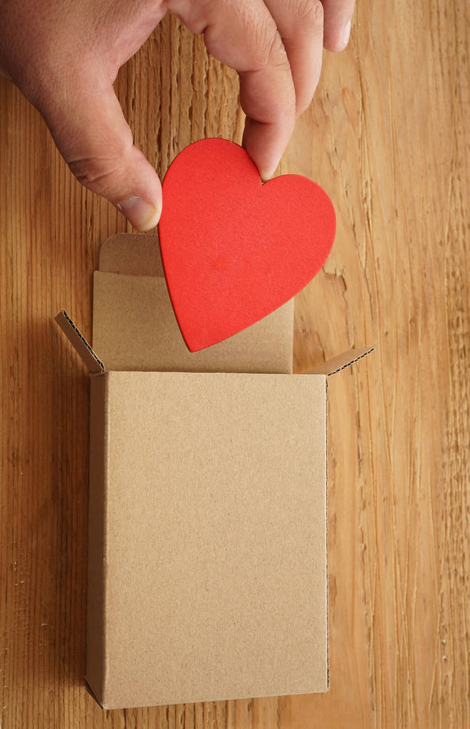 αρσενική χέρι κρατάει μία κόκκινη καρδιά πάνω από την ανοικτή συσκευασία δώρου σε ξύλινο υπόβαθρο. Happy Ημέρα του Αγίου Βαλεντίνου. 14 Φεβρουαρίου.    - Φωτογραφία, εικόνα