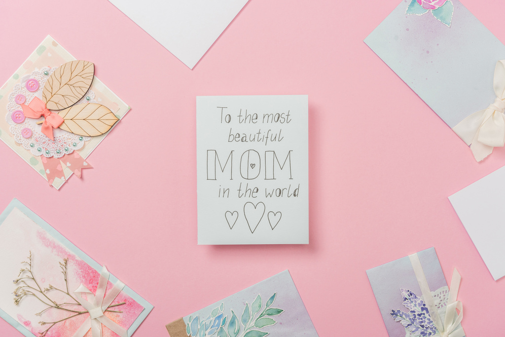 witte wenskaart voor Moederdag met "om de mooiste moeder in de wereld" handgeschreven tekst en harten en verschillende briefkaarten gerangschikt rond op roze achtergrond - Foto, afbeelding