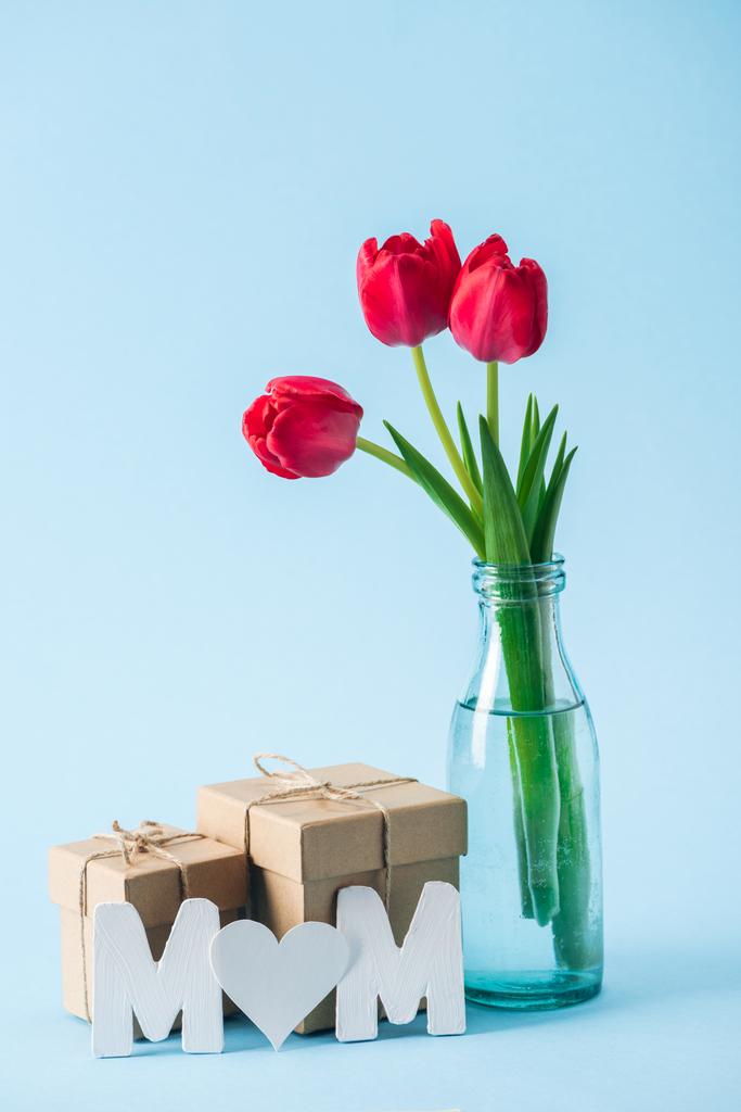 Geschenkboxen, Strauß roter Tulpen und Papierwort Mama mit herzförmigem Wurf "o" auf blauem Hintergrund - Foto, Bild