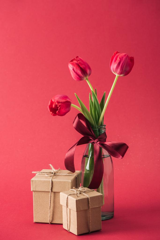 boeket van rode tulpen met rode satijnen boog en geschenk dozen op rode achtergrond - Foto, afbeelding
