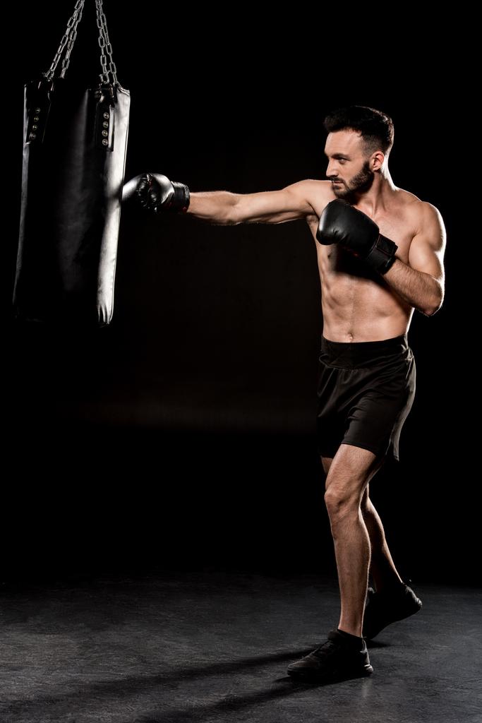 bärtiger Athlet mit Boxhandschuhen schlägt Boxsack auf schwarzem Hintergrund - Foto, Bild