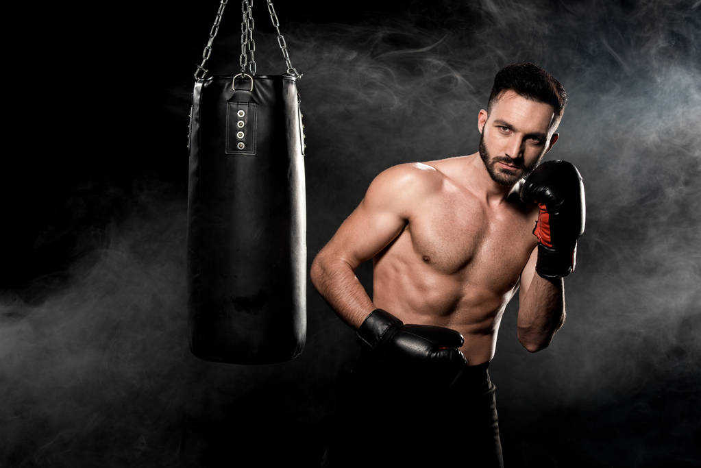 schöner Sportler in Boxhandschuhen, der neben Boxsack auf schwarz mit Rauch steht - Foto, Bild