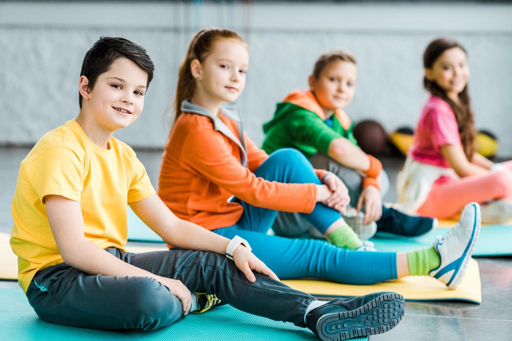 Crianças pré-adolescentes alegres sentadas em tapetes no ginásio
 - Foto, Imagem