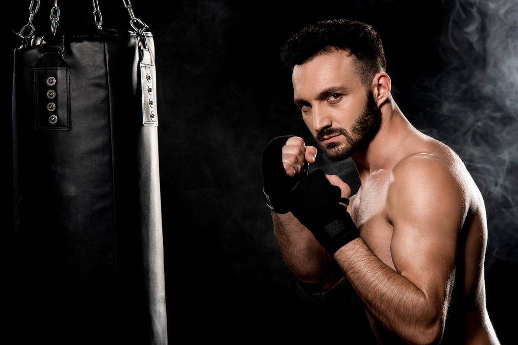 mięśni fighter stojąc w pozie boks w pobliżu worek treningowy na czarnym tle - Zdjęcie, obraz