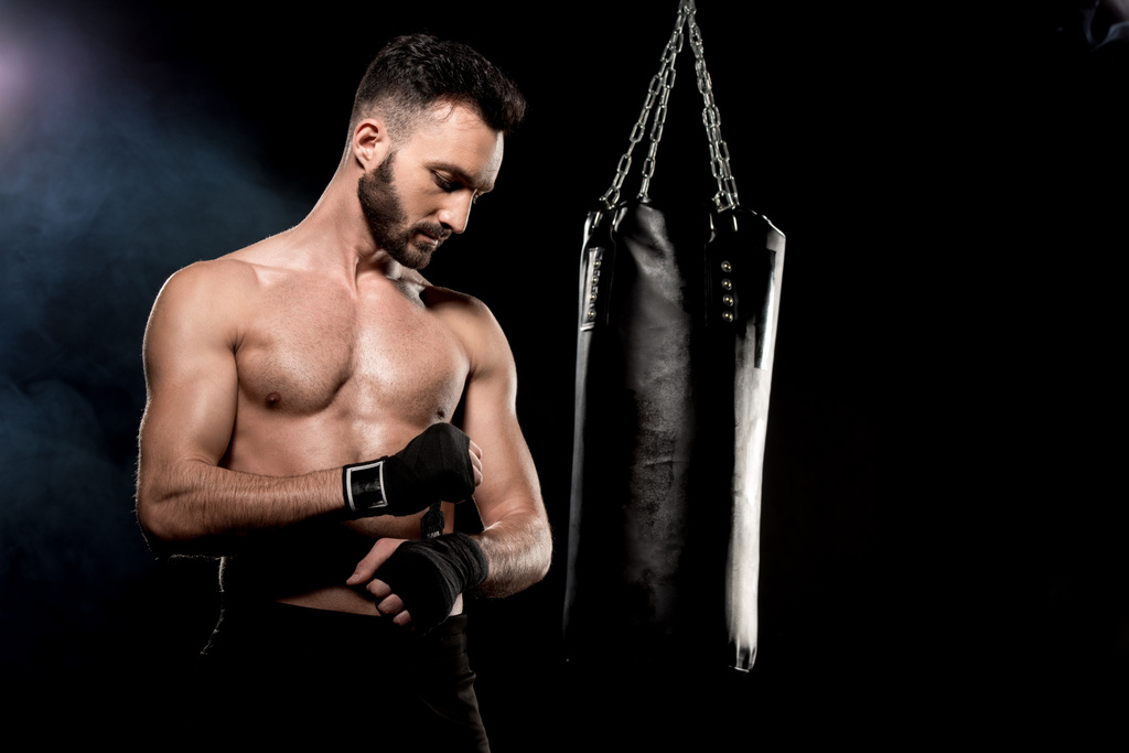 handsome athletic boxer wrapping up gloves near punching bag on black background - Photo, Image