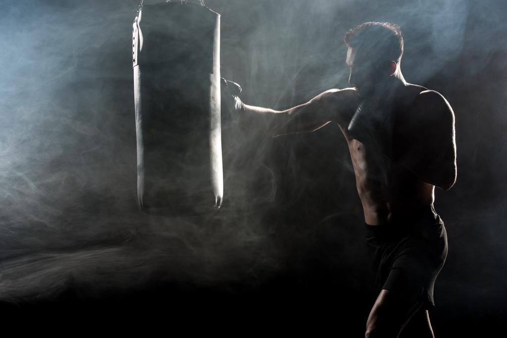 silhouette of sportsman in boxing gloves hitting punching bag on black with smoke - Photo, Image