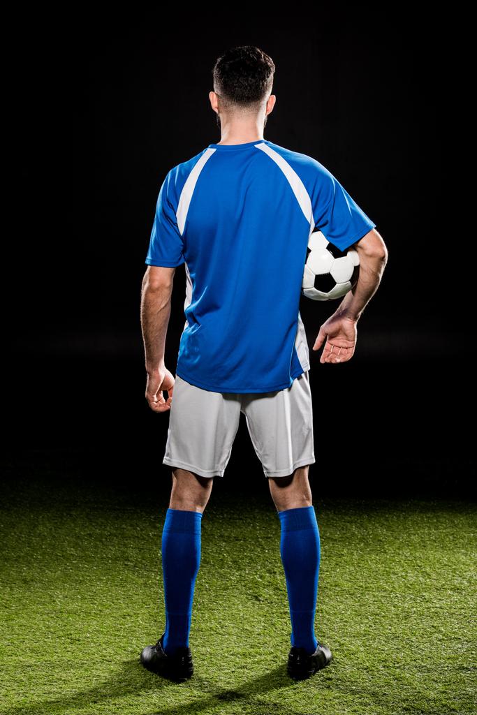 vista trasera del deportista sosteniendo la pelota y de pie sobre hierba aislado en negro
 - Foto, imagen