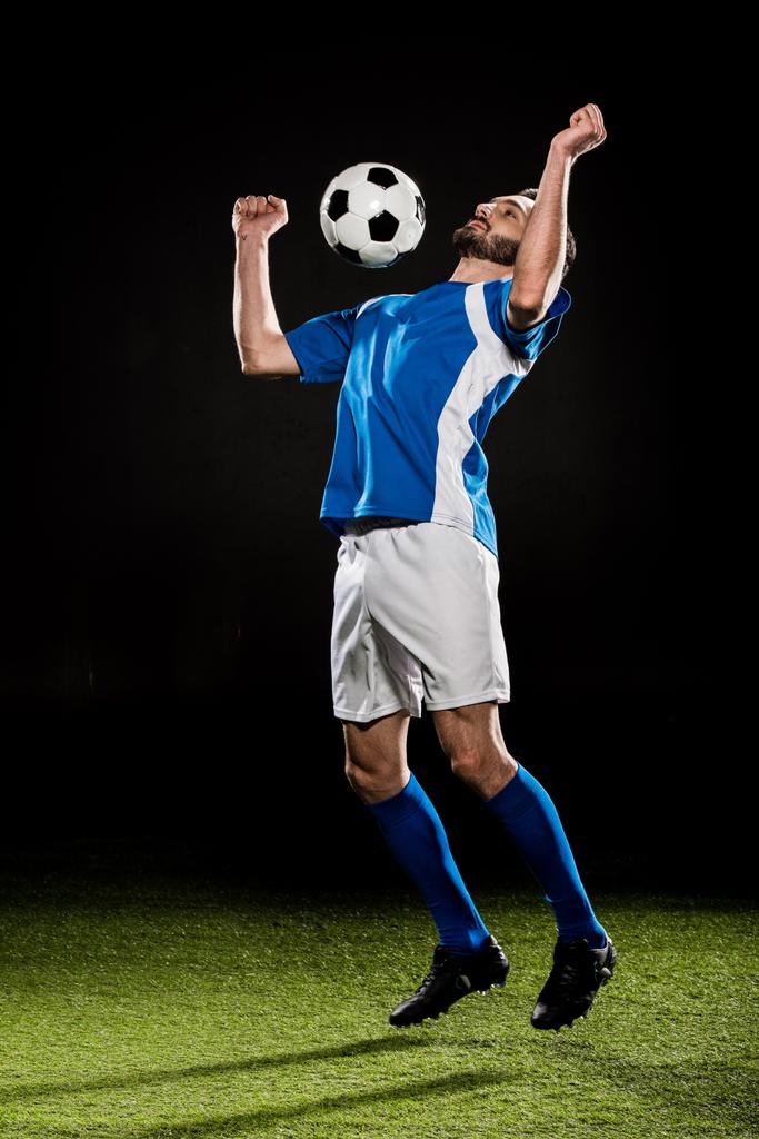schöner Fußballer in Uniform springt mit Ball isoliert auf schwarz - Foto, Bild