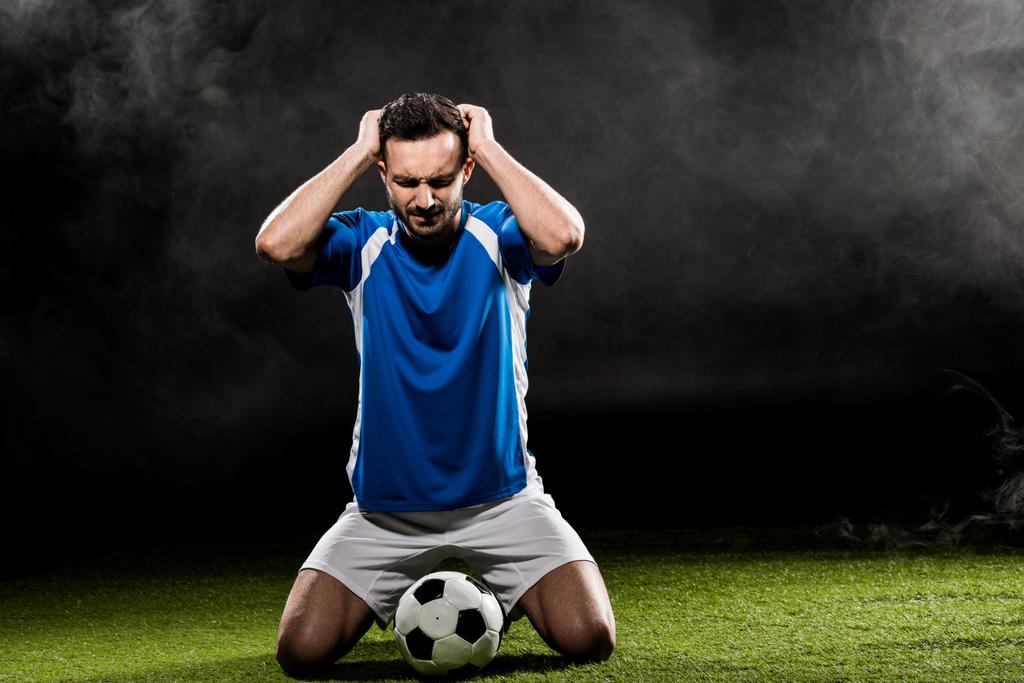 handsome football player holding head while sitting on grass on black with smoke - Photo, Image