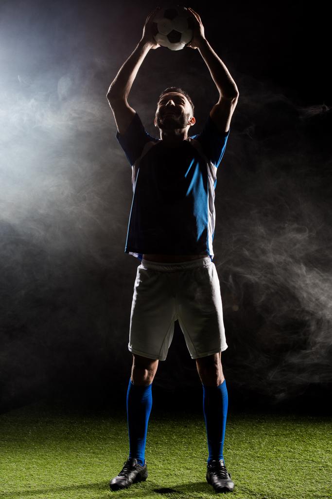 煙と黒地の頭の上にボールを保持しているフットボール選手のシルエット   - 写真・画像