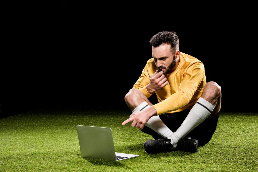 árbitro bonito soprando no apito e apontando com o dedo para laptop enquanto sentado na grama isolada no preto
 - Foto, Imagem