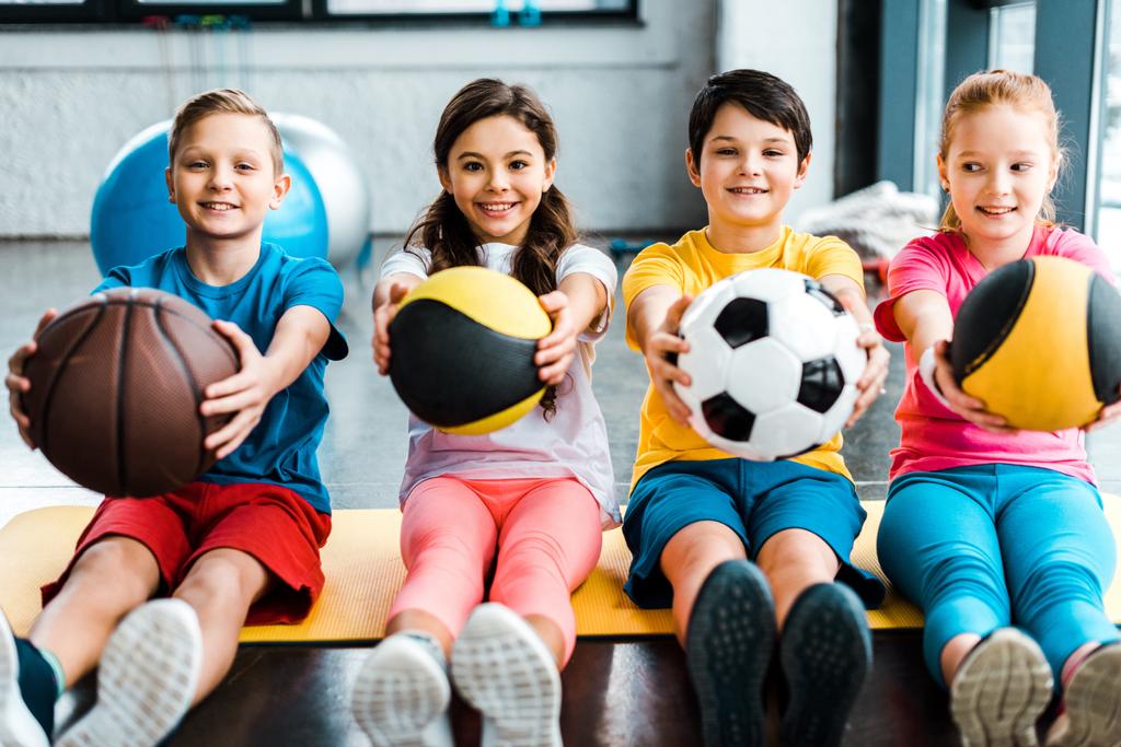 Fröhliche Kinder sitzen auf Fitnessmatte mit Bällen - Foto, Bild