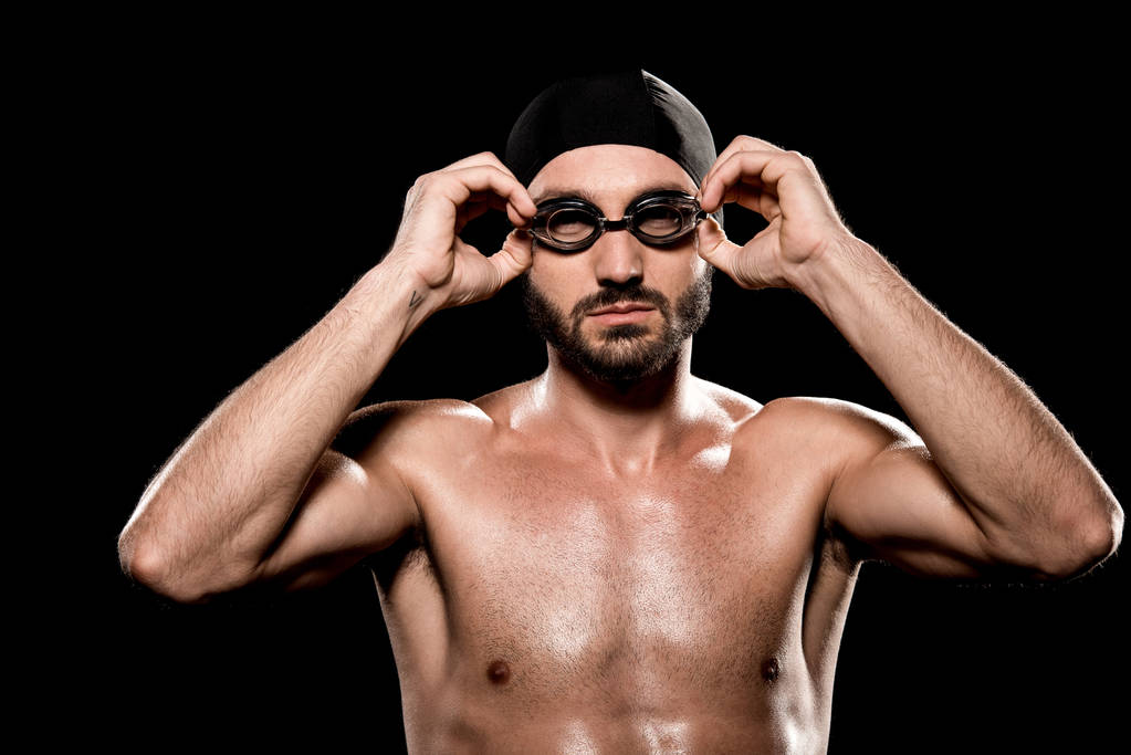 confident swimmer standing in swimming cap and touching goggles isolated on black  - Photo, Image
