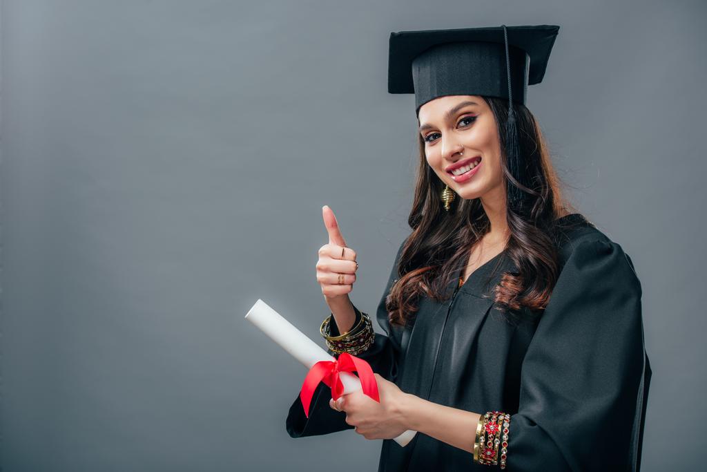 Улыбающаяся индийская студентка в академическом платье и шляпе выпускного, держащая диплом и показывая большой палец вверх, изолированный на сером
 - Фото, изображение