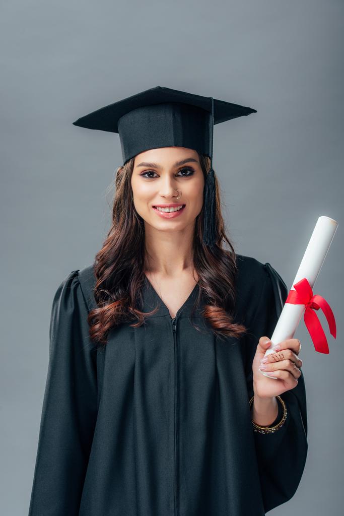 lächelnde indische Studentin in akademischem Gewand und Diplomhut, isoliert auf grau - Foto, Bild