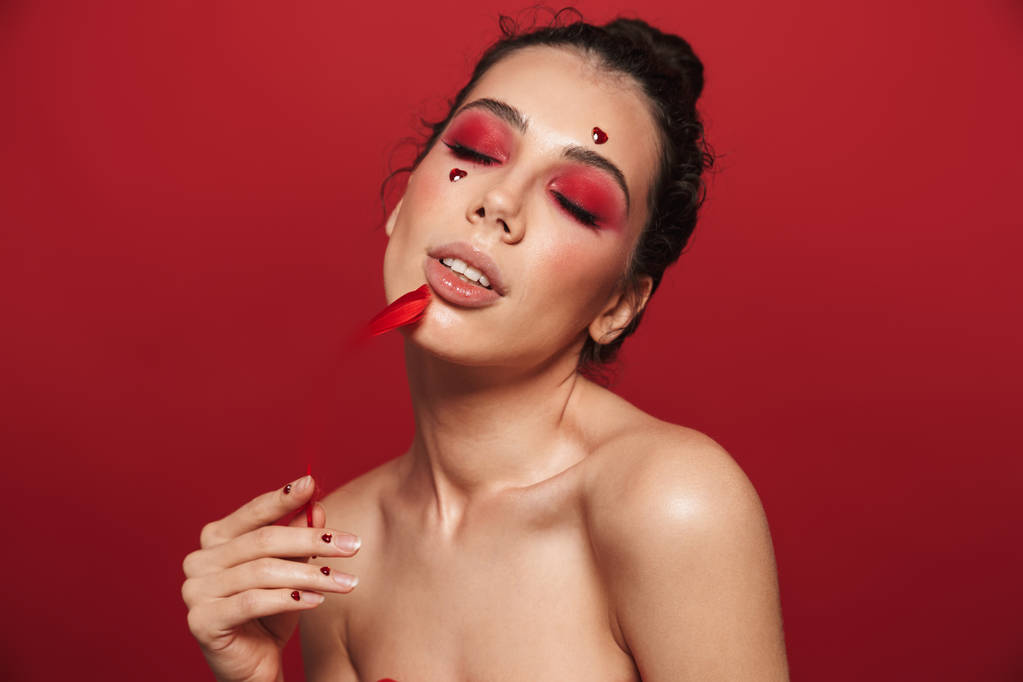 Portret van de schoonheid van een aantrekkelijke jonge topless vrouw dragen make-up staande geïsoleerd op rode achtergrond, poseren, met een veer - Foto, afbeelding