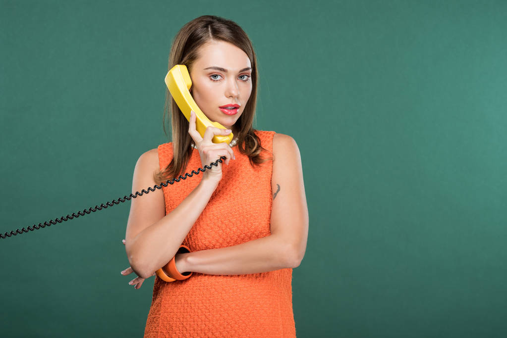 beautiful stylish woman looking at camera and talking on retro telephone isolated on green with copy space - Photo, Image