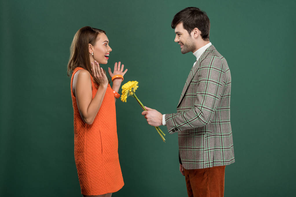 hombre guapo en ropa vintage presentando flores a hermosa mujer feliz aislado en verde
 - Foto, Imagen