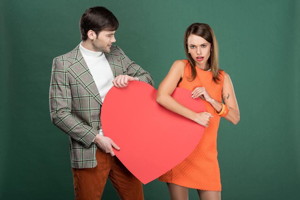 Mann schaut unzufriedene Frau mit herzförmiger Papierkarte auf grünem Grund an - Foto, Bild