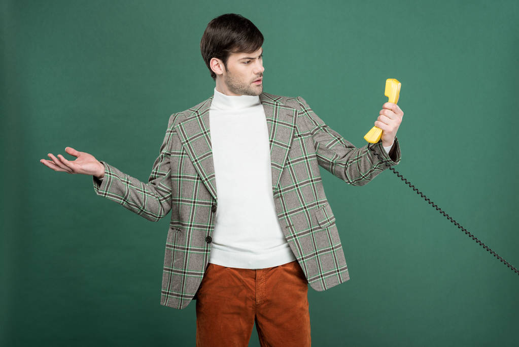 Überraschter gutaussehender Mann in Vintage-Klamotten blickt auf Retro-Telefon und gestikuliert mit der Hand isoliert auf Grün - Foto, Bild