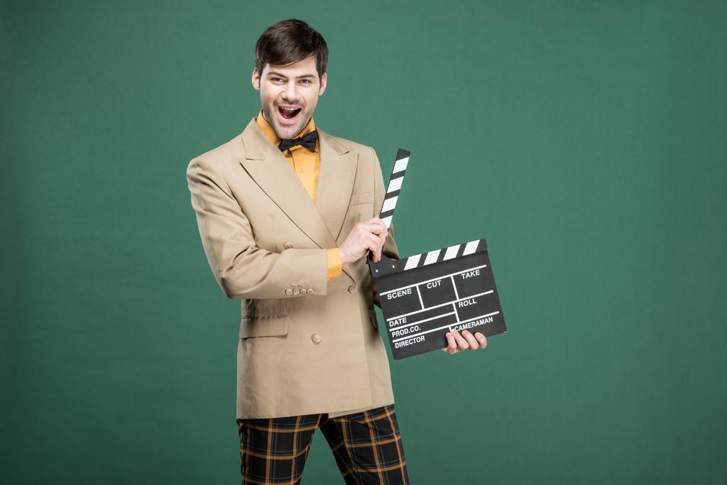 excited handsome man in vintage clothes looking at camera and holding film clapperboard isolated on green - Photo, Image