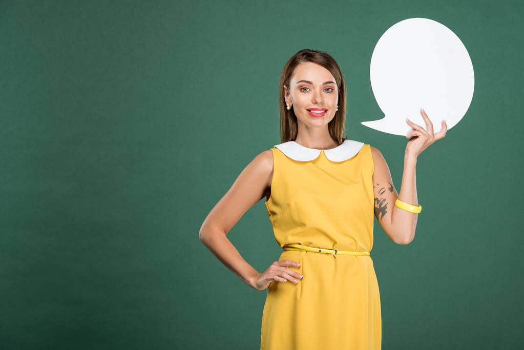 beautiful smiling woman in yellow dress looking at camera and holding speech bubble isolated on green - Photo, Image