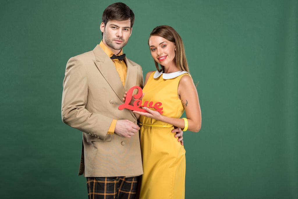 beautiful couple in vintage clothes looking at camera and holding decorative word 'love' isolated on green - Photo, Image