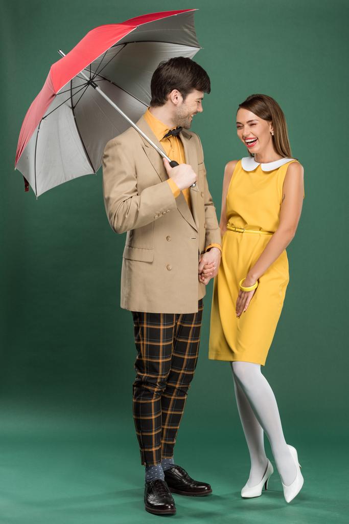 mooi lachen paar in vintage kleding poseren met paraplu op groene achtergrond - Foto, afbeelding