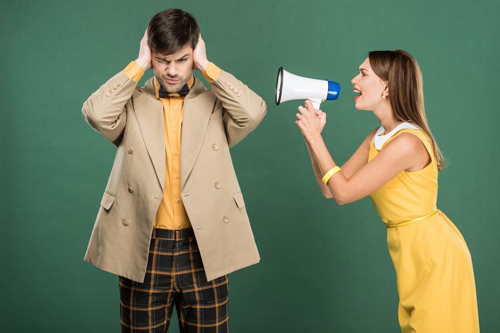 man in vintage clothes covering ears with hands while angry woman yelling in loudspeaker isolated on green - Photo, Image