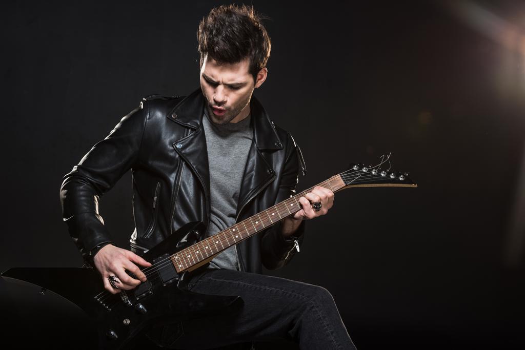 όμορφος rocker σε δερμάτινο μπουφάν παίζοντας ηλεκτρική κιθάρα σε μαύρο φόντο - Φωτογραφία, εικόνα