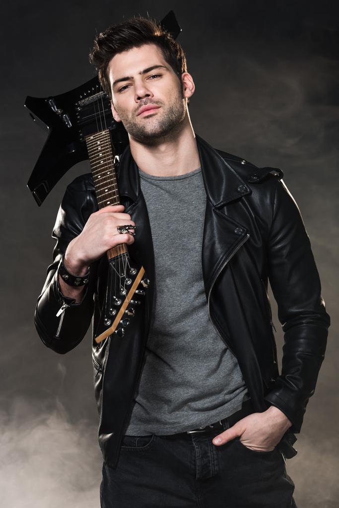 handsome rocker holding electric guitar and looking at camera on dark smoky background - Photo, Image