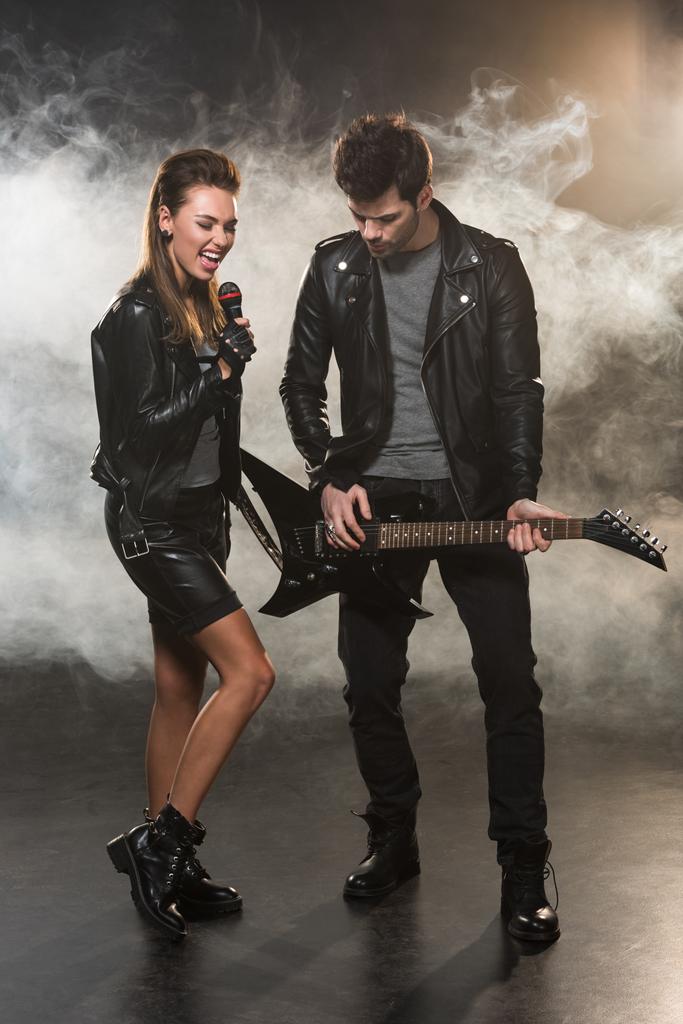 schöne Frau in Lederjacke singt, während Mann auf verrauchtem Hintergrund E-Gitarre spielt - Foto, Bild