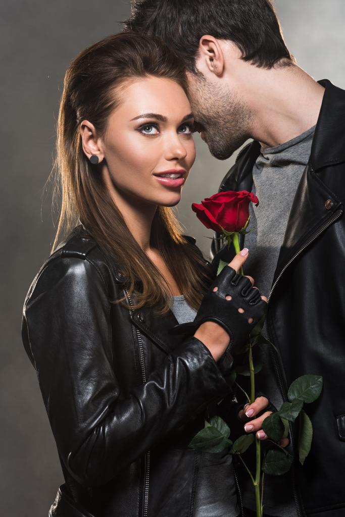 beautiful couple in leather jackets posing with red rose on grey background - Photo, Image