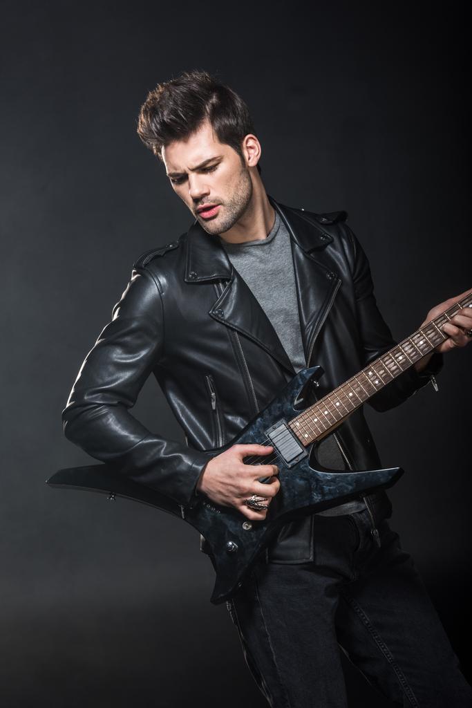 handsome rocker in leather jacket playing electric guitar isolated on black - Photo, Image