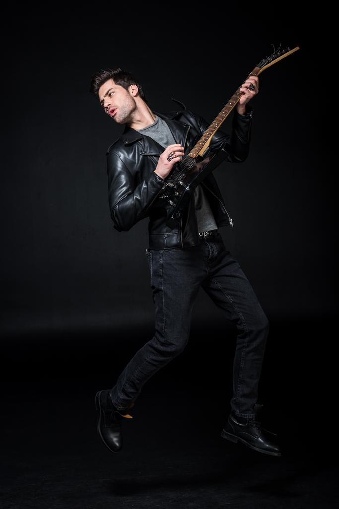 przystojny rocker w skórzanej kurtce grać na gitarze elektrycznej na czarnym tle - Zdjęcie, obraz