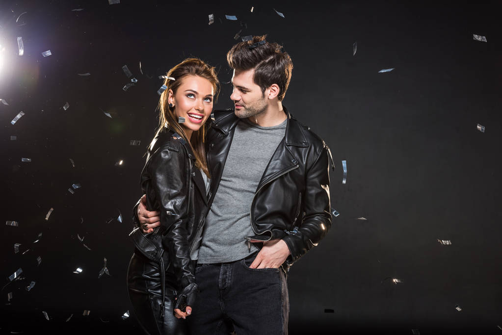 beautiful couple in leather jackets hugging with falling confetti on black background - Photo, Image