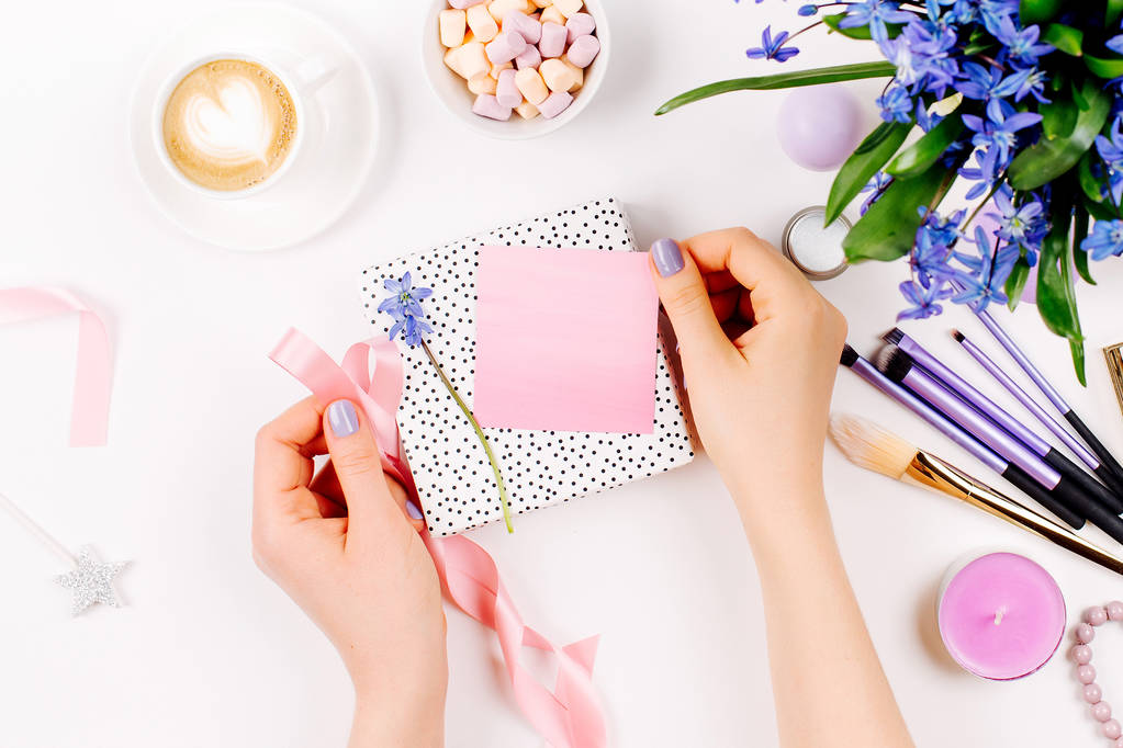 Une femme déballonne un cadeau sur fond de bouquet de printemps bleu, tasse à café, bonbons et accessoires cosmétiques. Pose plate de style
 - Photo, image