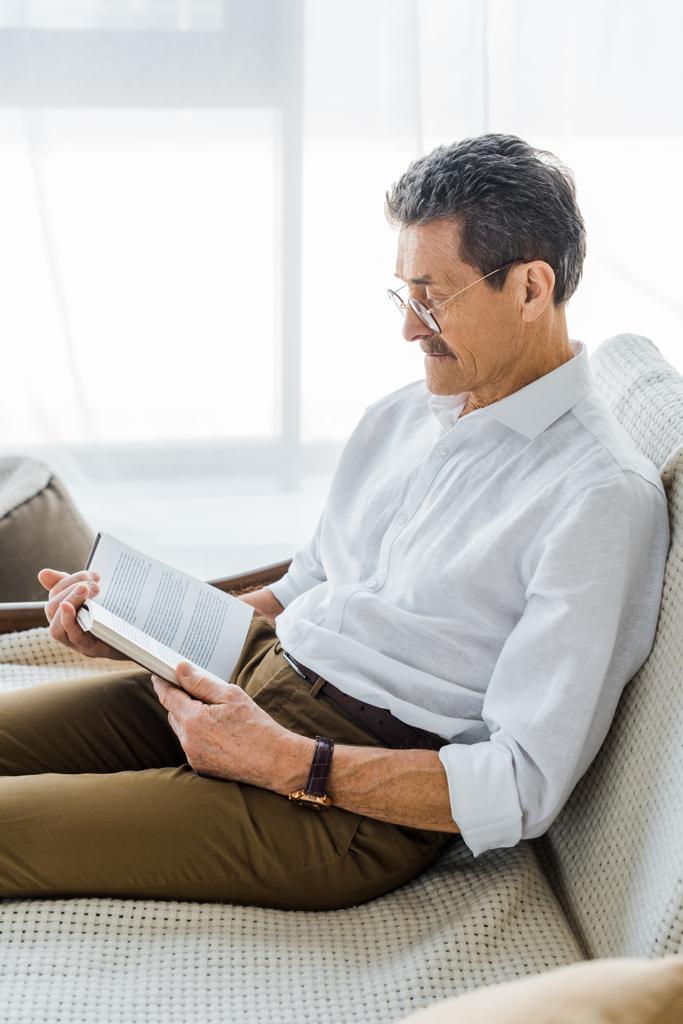  Senior uomo lettura libro mentre seduto sul divano a casa
 - Foto, immagini
