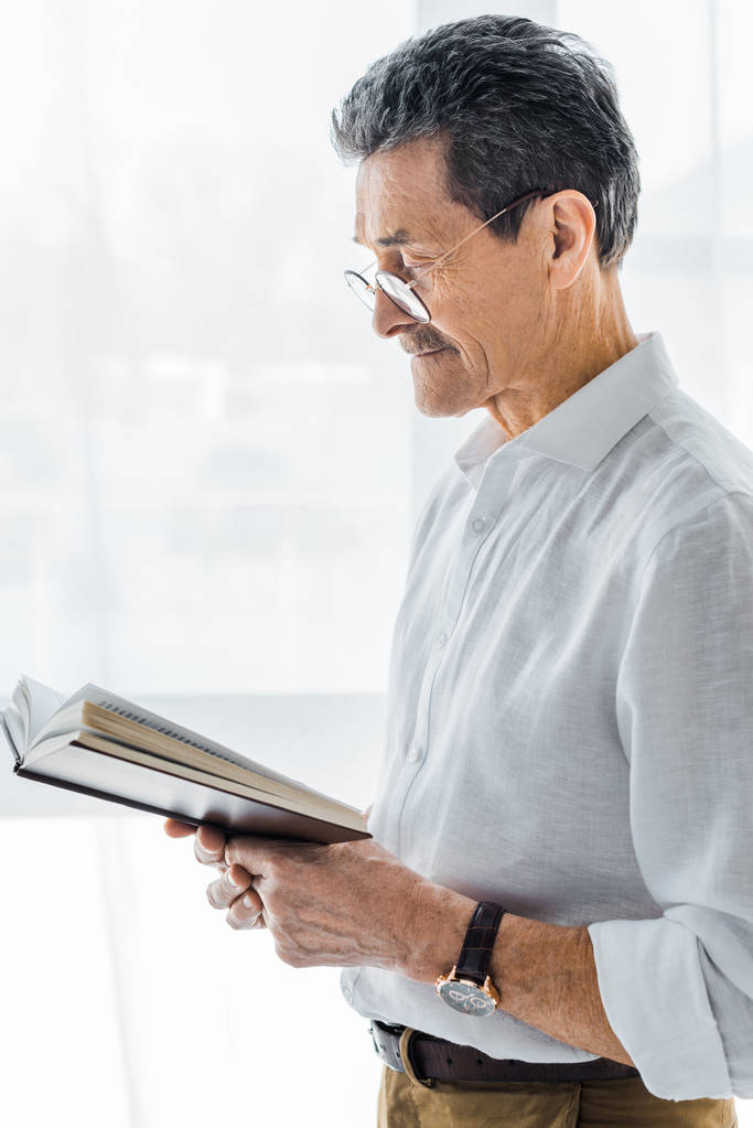 пенсионер в очках стоя и читая книгу дома
 - Фото, изображение