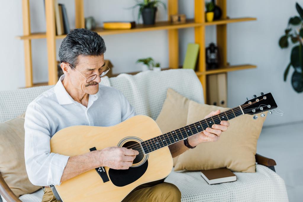 šťastný důchodce v brýlích hraje na akustickou kytaru doma - Fotografie, Obrázek