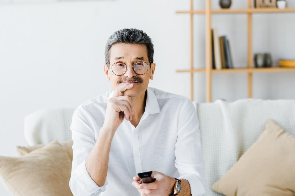 cheerful senior man with mustache sitting on sofa and holding remote control - Photo, Image