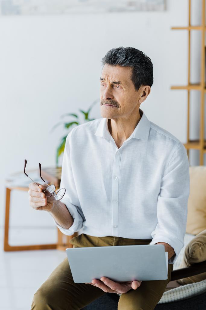 pensionista pensativo com bigode segurando óculos e laptop em casa
 - Foto, Imagem
